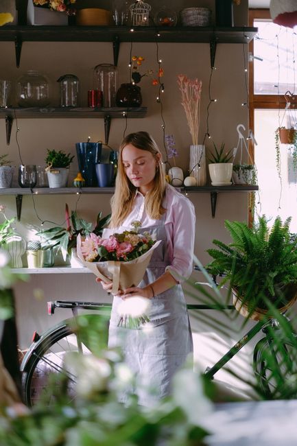 A young woman with long blonde hair, dressed in a white blouse and apron, is holding a bouquet of fresh flowers wrapped in light brown paper. The bouquet features pink roses, white lilies, and greener